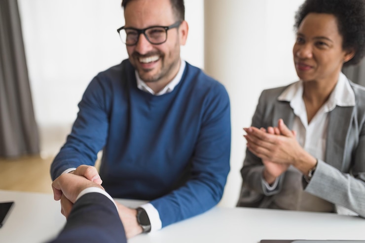 A smiling man in a blue sweater shaking hands with a client while a colleague applauds